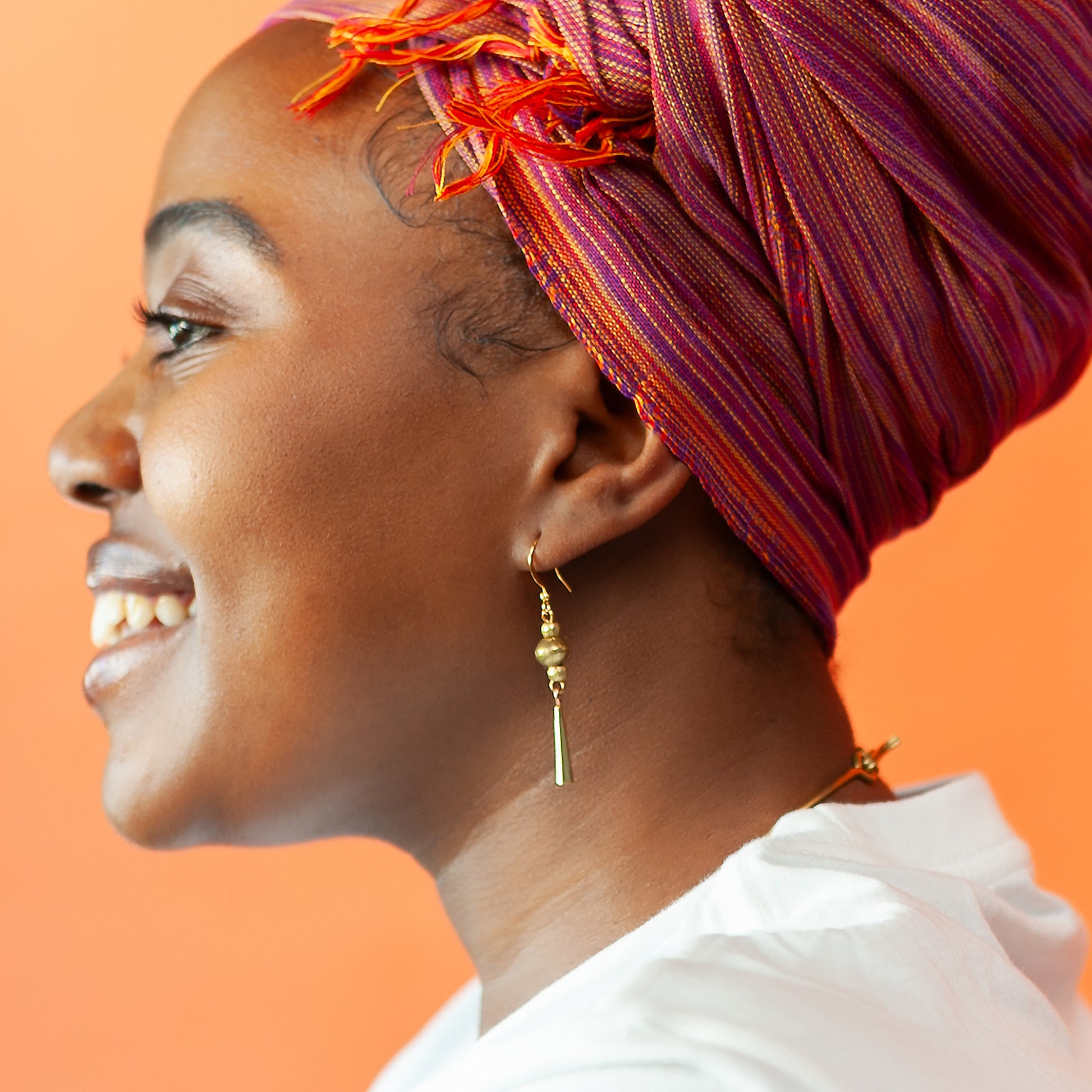 A close-up side profile of a smiling woman wearing a vibrant magenta and orange striped headwrap and a gold dangle hoop earring, set against a solid orange background.