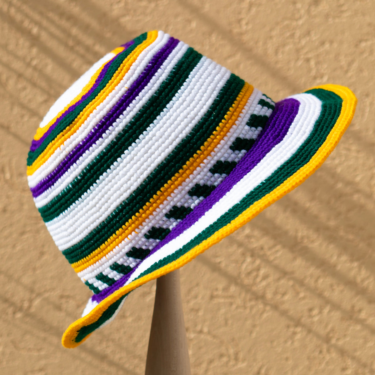 A crocheted bucket hat with horizontal stripes of green, white, purple, and yellow, displayed on a wooden stand against a textured tan background.