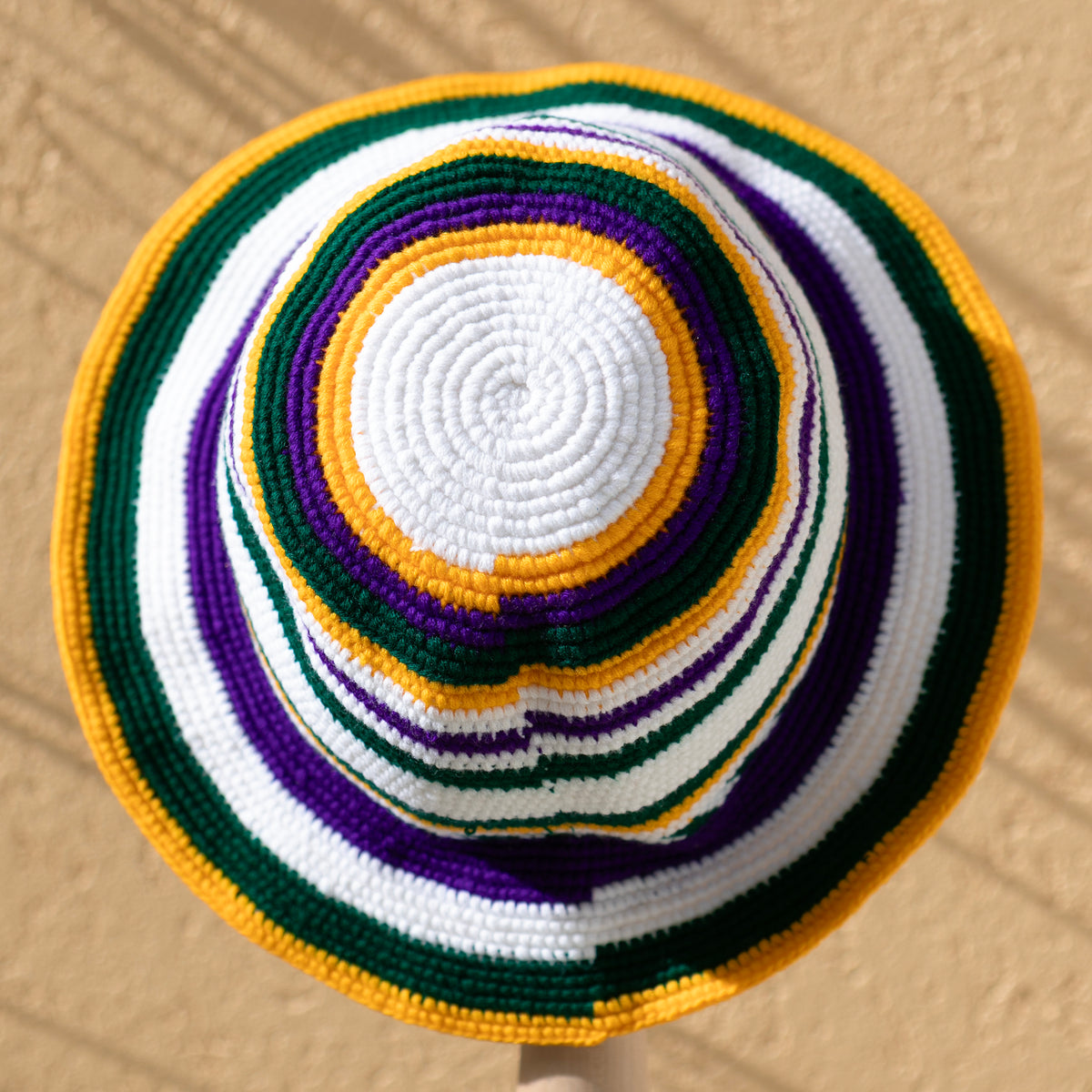 A crocheted sun hat with concentric stripes of white, purple, green, and yellow, photographed from directly above against a textured beige wall.