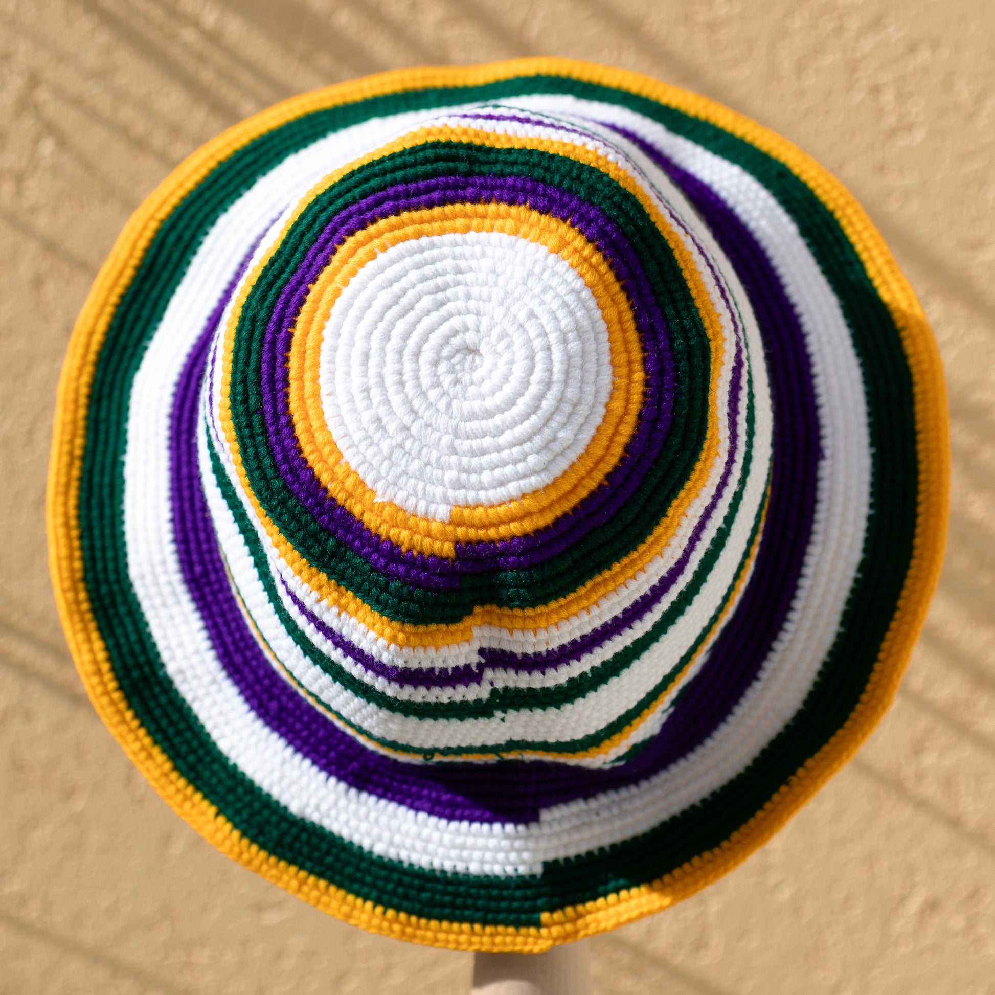 A crocheted sun hat with concentric stripes of white, purple, green, and yellow, photographed from directly above against a textured beige wall.