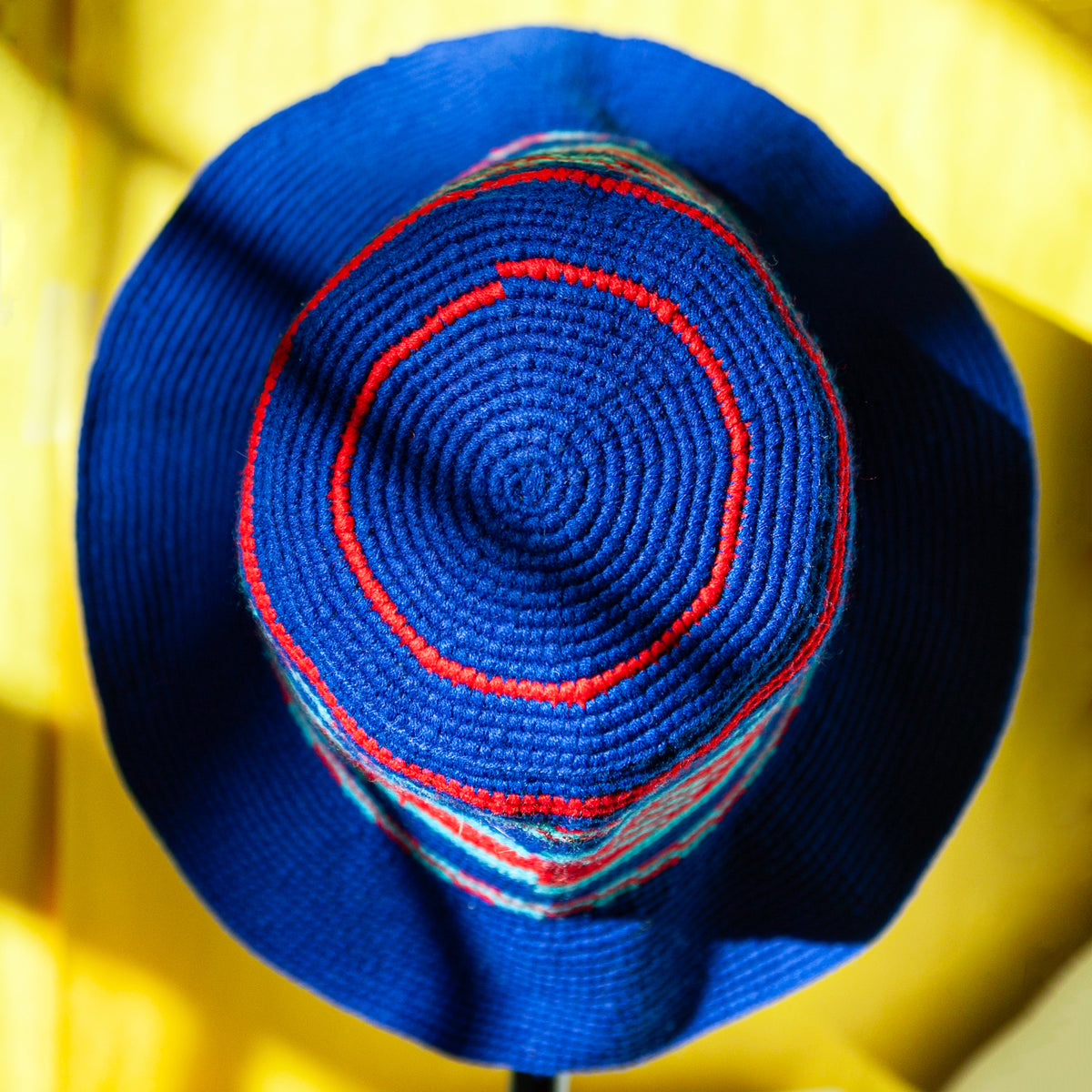 A close-up top-down view of a blue knit hat with concentric red and white stripes on the crown, set against a bright yellow background.