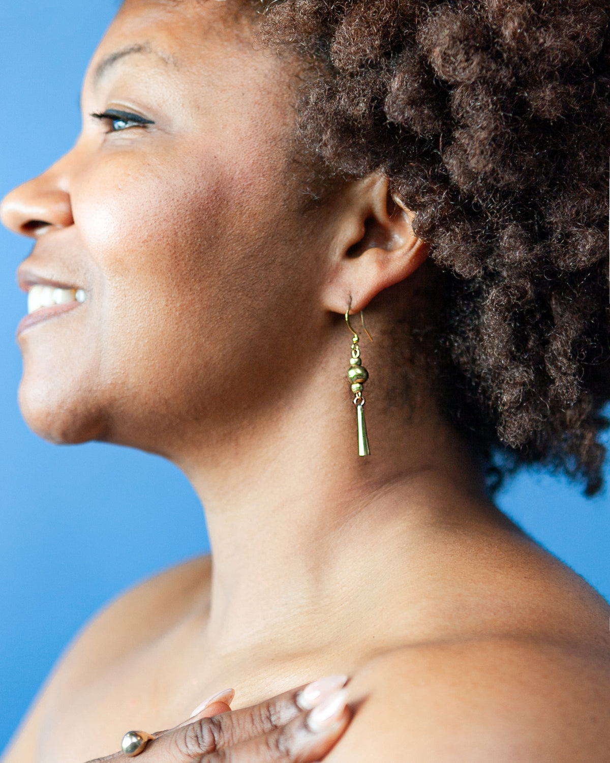 A close-up side profile of a woman with curly dark hair wearing a single dangling gold earring against a solid blue background.