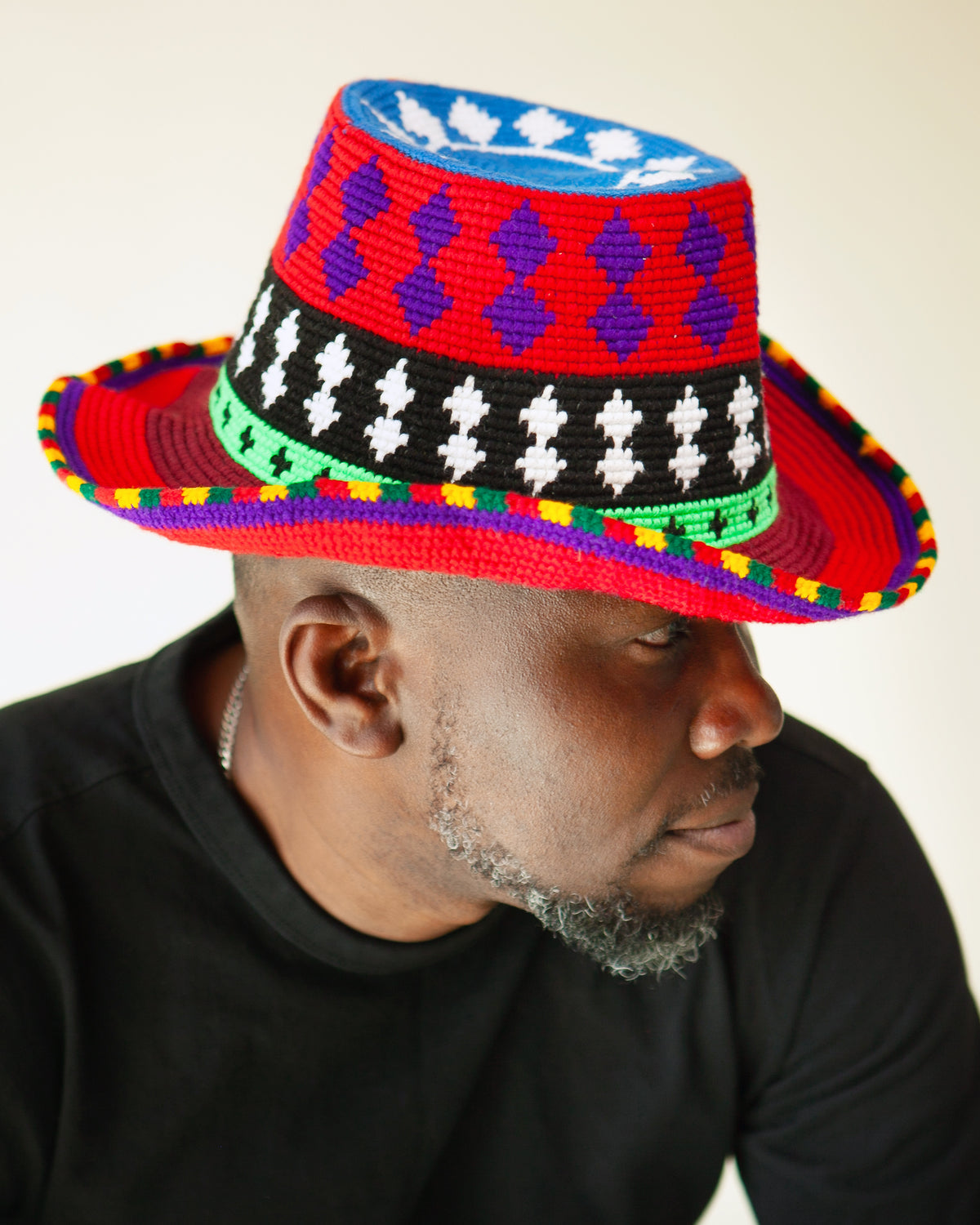A man wearing a vibrant, hand-beaded African-style fedora hat with red, blue, black, white, green, and yellow patterns, photographed against a plain light background.